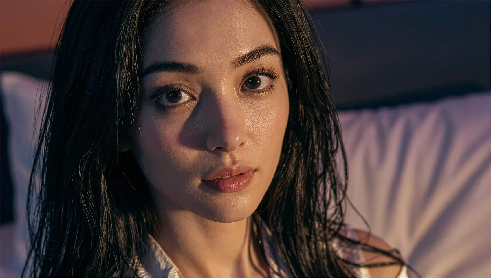Wide close frame of a fictional AI character with wet dark hair lying on a pillow in warm pink light